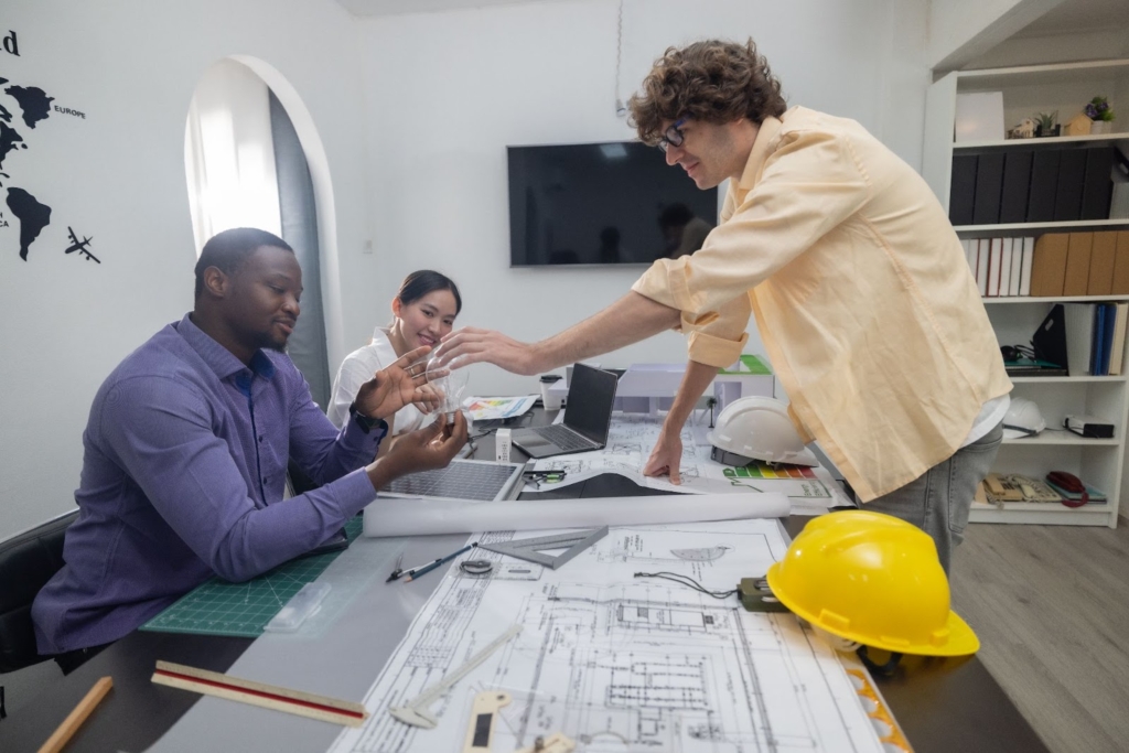 A male consultant wearing a powder blue long-sleeved polo shirt and dark-rimmed glasses is seated at a table, discussing project cost estimations with three business owners.