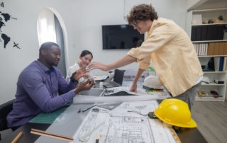 A male consultant wearing a powder blue long-sleeved polo shirt and dark-rimmed glasses is seated at a table, discussing project cost estimations with three business owners.