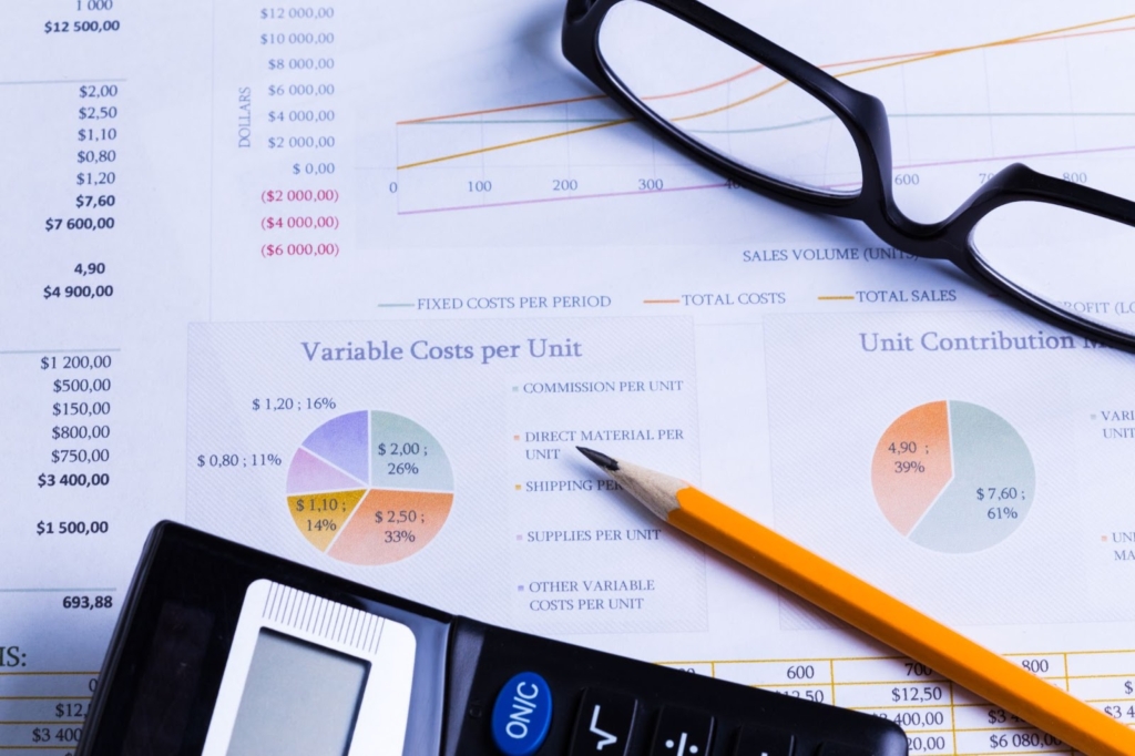 A black calculator, black-framed eyeglasses, and a pencil rest on sheets filled with manual project cost calculations, showcasing the task's complexity and tedious nature.