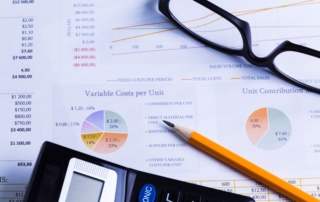 A black calculator, black-framed eyeglasses, and a pencil rest on sheets filled with manual project cost calculations, showcasing the task's complexity and tedious nature.