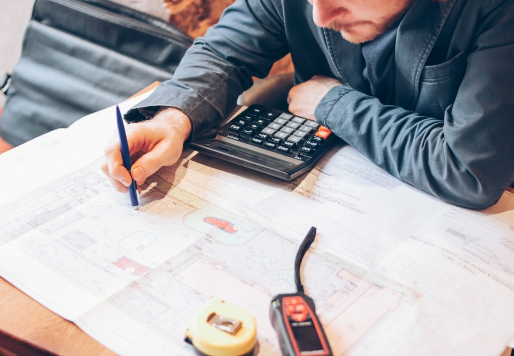 A male architect in a blue-gray jacket reviewing blueprints at a cafe table, with a satchel by his side, calculating cost estimates for a residential restoration project.