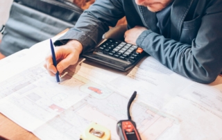 A male architect in a blue-gray jacket reviewing blueprints at a cafe table, with a satchel by his side, calculating cost estimates for a residential restoration project.