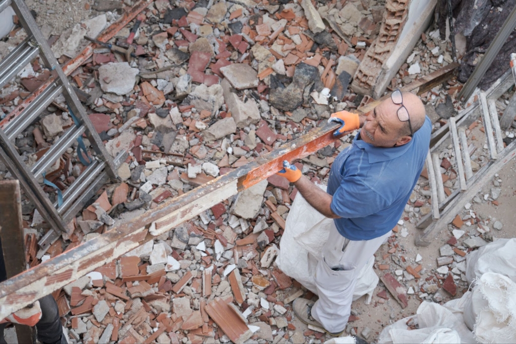 A bald handyman wearing a blue polo shirt and eyeglasses on his head stands on rubble while removing old metal beams in a fully demolished house. 