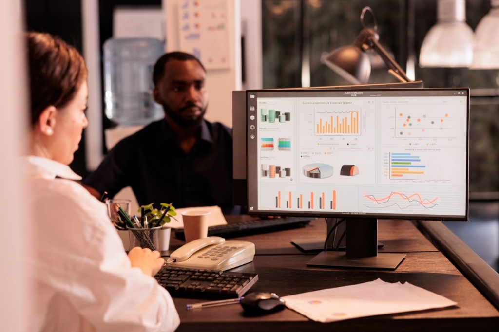 A female executive in a cream-colored suit analyzes marketing research statistics using computer software with a male colleague seated across from her.