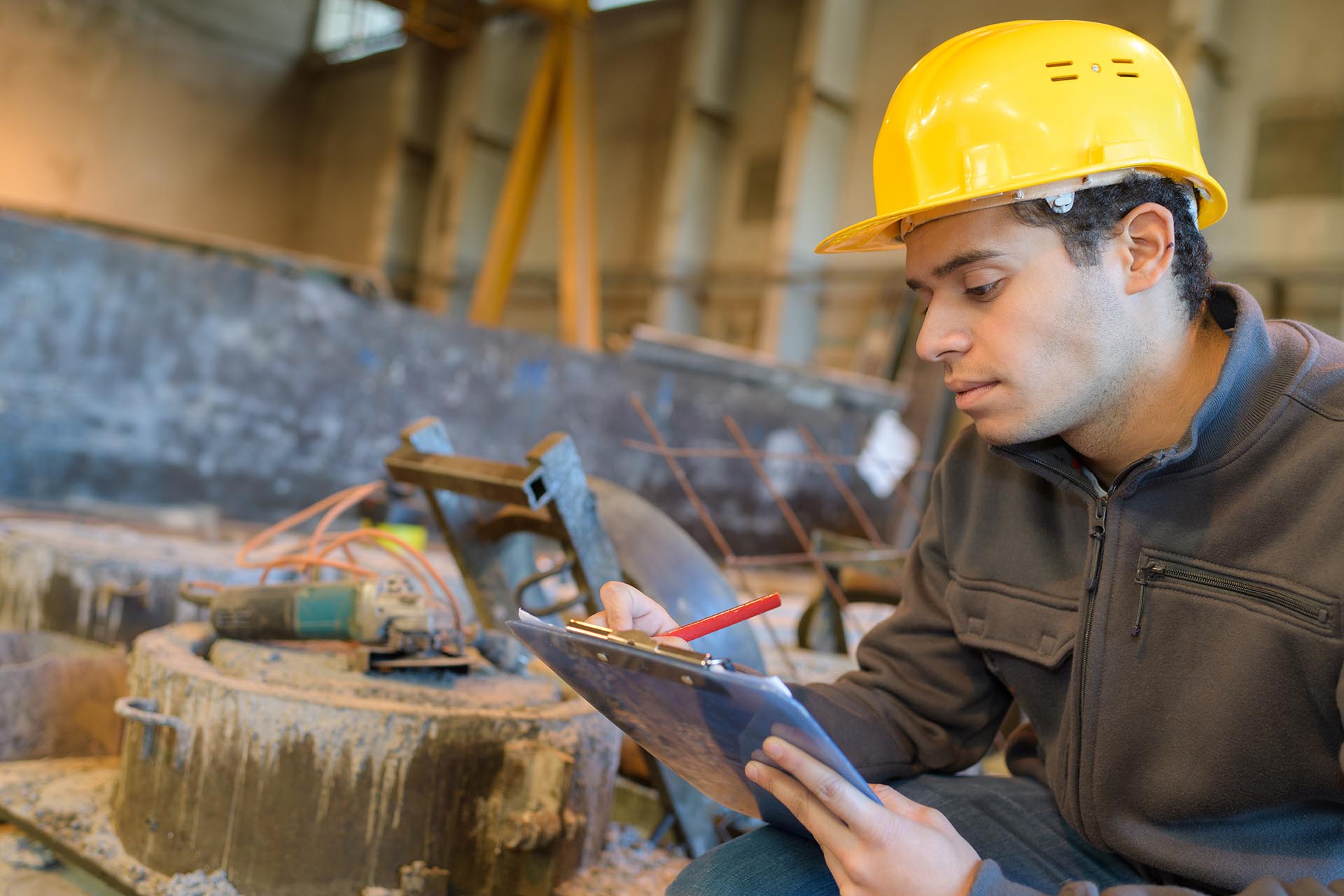 A man in a hard hat writing on a clipboard at a construction site.