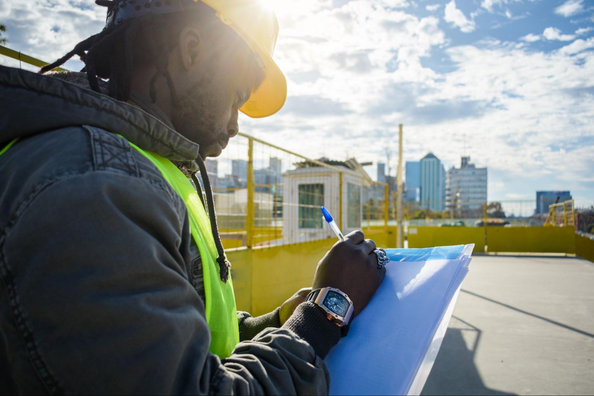 A rear view of a male engineer in the field making notes on paper, with the commercial district and skyline in the background, focusing on roofing estimates.