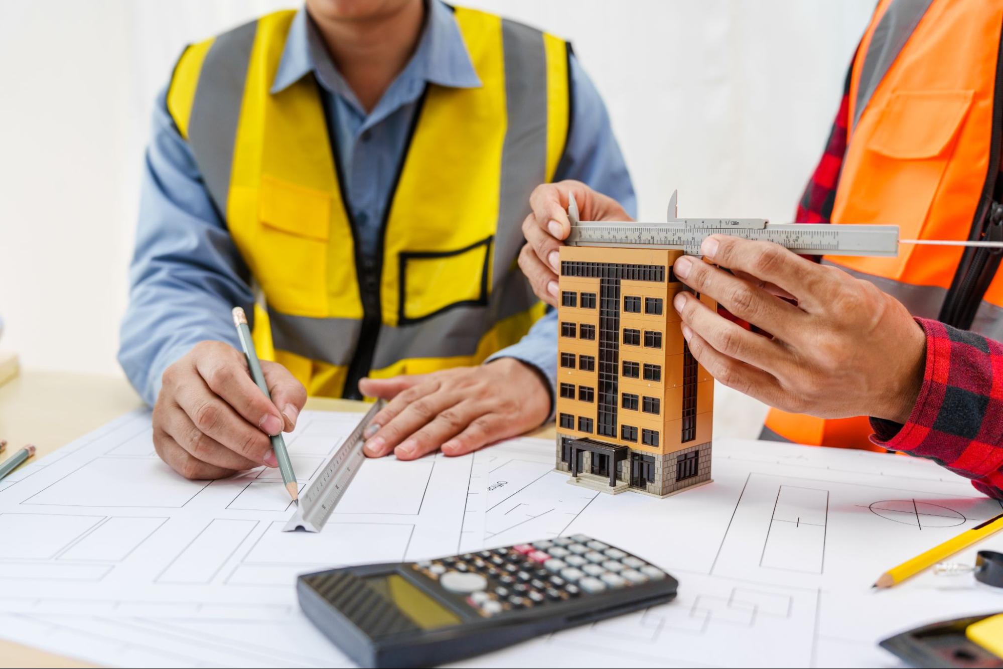 Two unidentified male construction contractors are wearing different-colored safety vests. One is drawing, while the other measures a scale building model to discuss structural plans and budget planning.