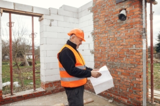 A senior male consultant in a hardhat and orange safety vest checking blueprints inside a devastated building, assessing storm damage.