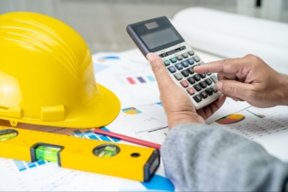 A close-up of an engineer's hand using a calculator with architectural blueprints, graphs, and a yellow helmet on the table, illustrating cost estimation and budget planning in construction.