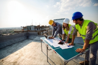 Three roofing contractors in safety vests and helmets review the building plan and discuss roofing estimates at the open rooftop.