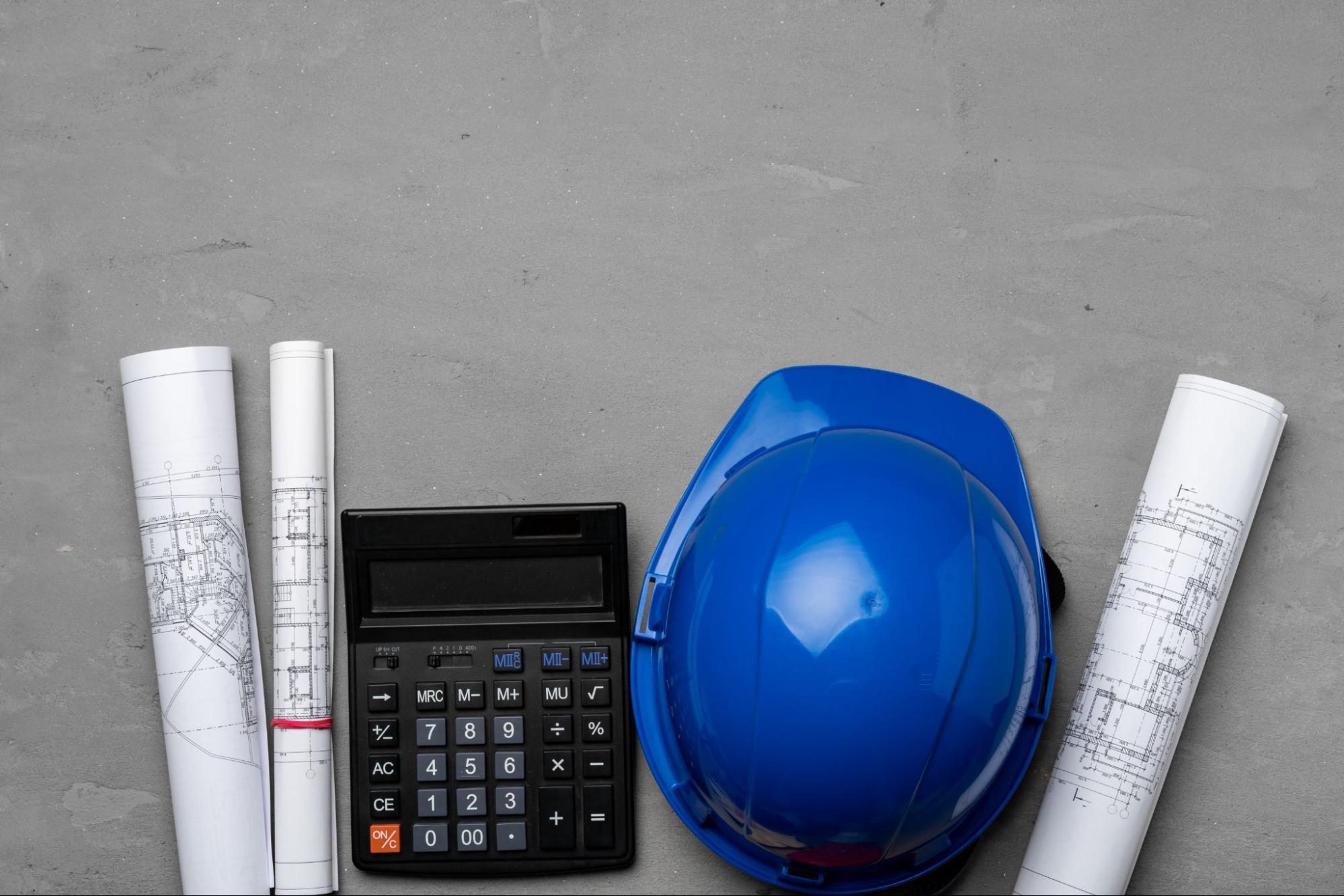 Top view of architect tools, including a blue hard hat, blueprints, and a calculator, illustrating cost estimation and budget planning.