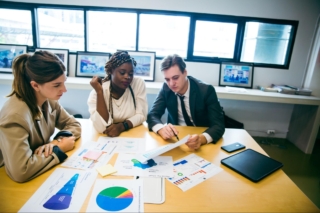 Two women and a man looking at graphs and charts in an office while sitting at a table.