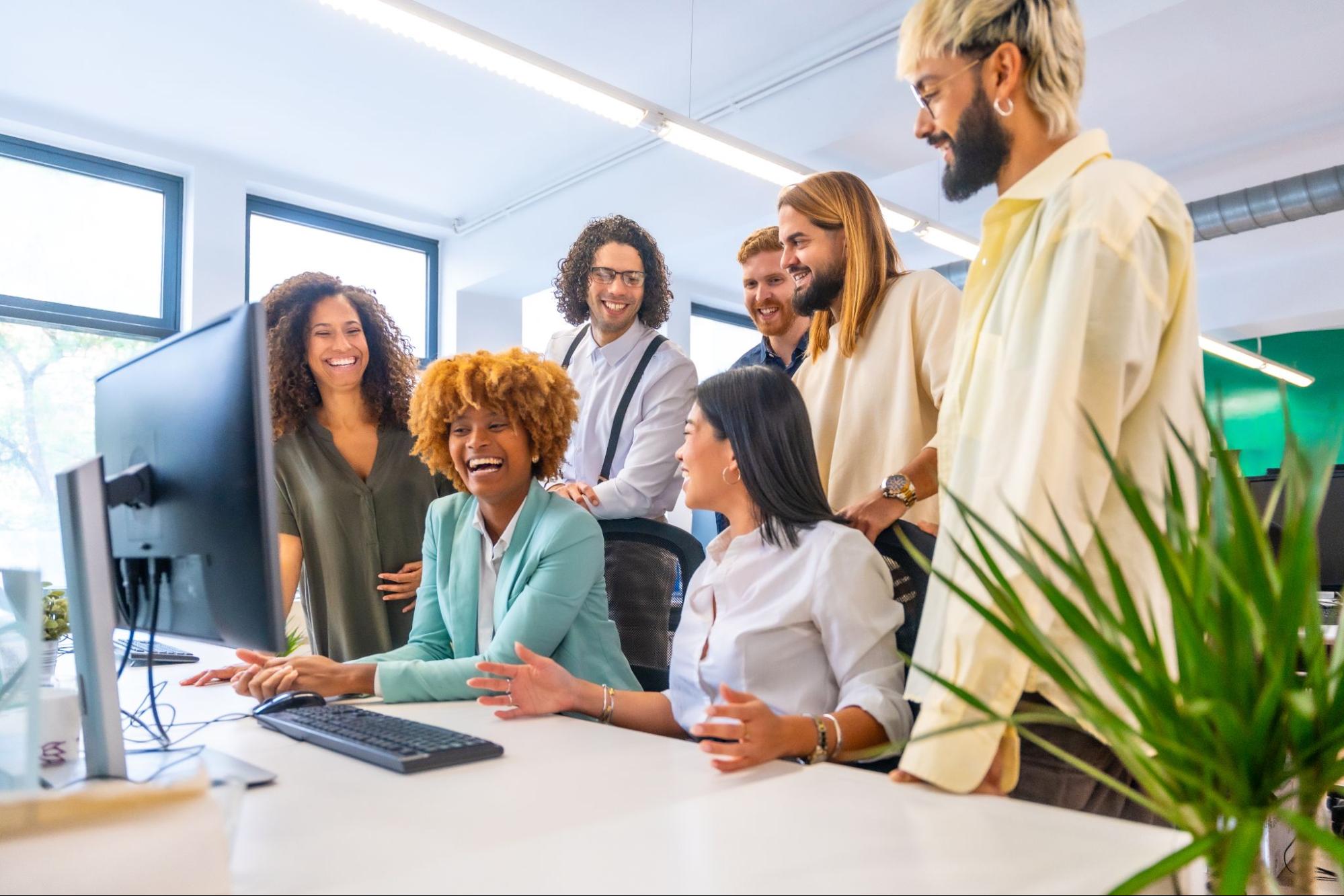 Group of employees standing and sitting around a computer monitor and smiling. 