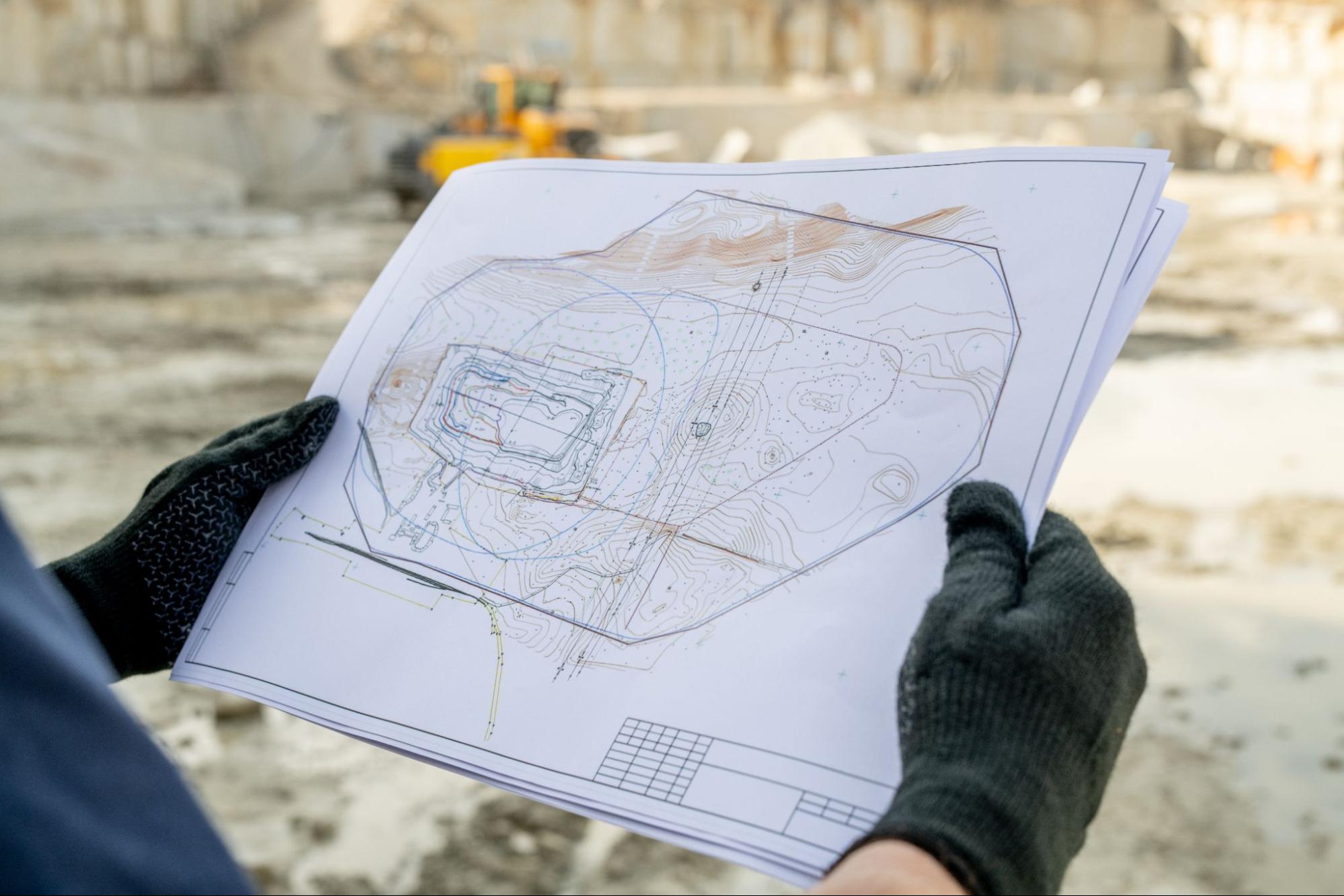 A person in gloves holding a blueprint on a construction site. 