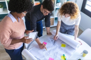 Three people in formal attire looking at blueprints.