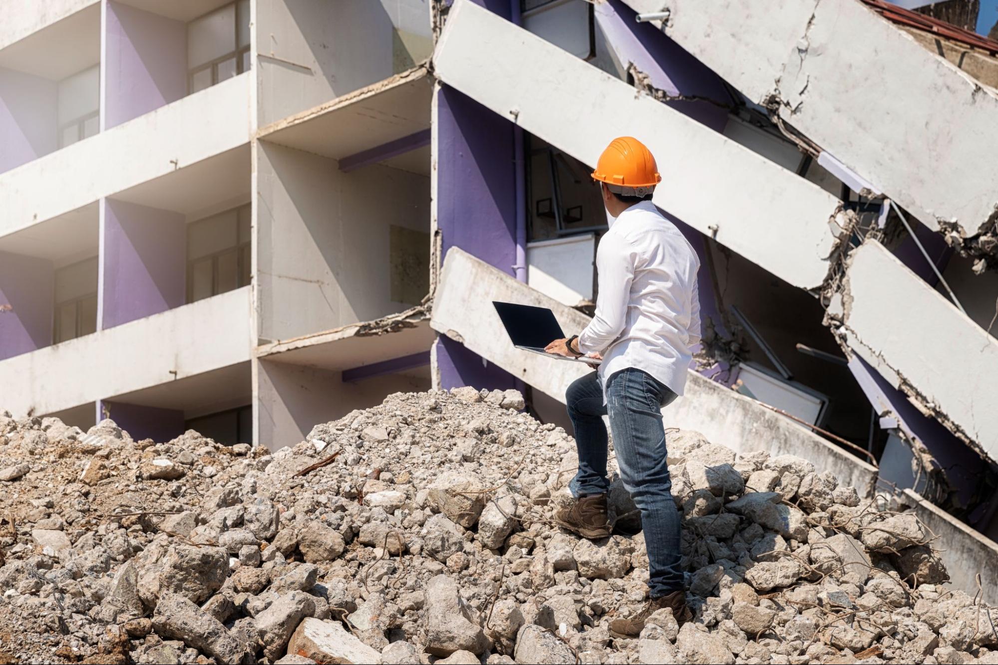 An engineer holding a laptop inspecting a collapsed building for damage.