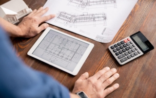 Man’s hands on a desk next to a tablet, calculator, and blueprint.