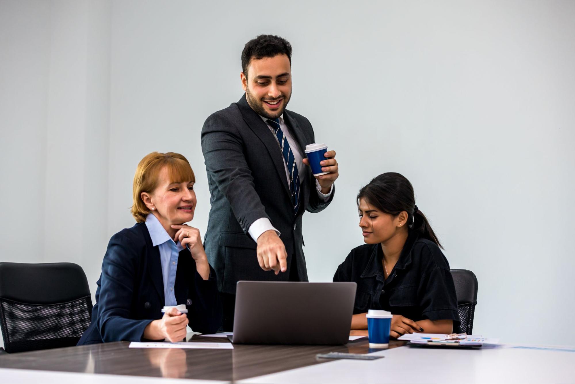 A male colleague gestures his hand to a laptop screen, discussing a topic with two female colleagues.