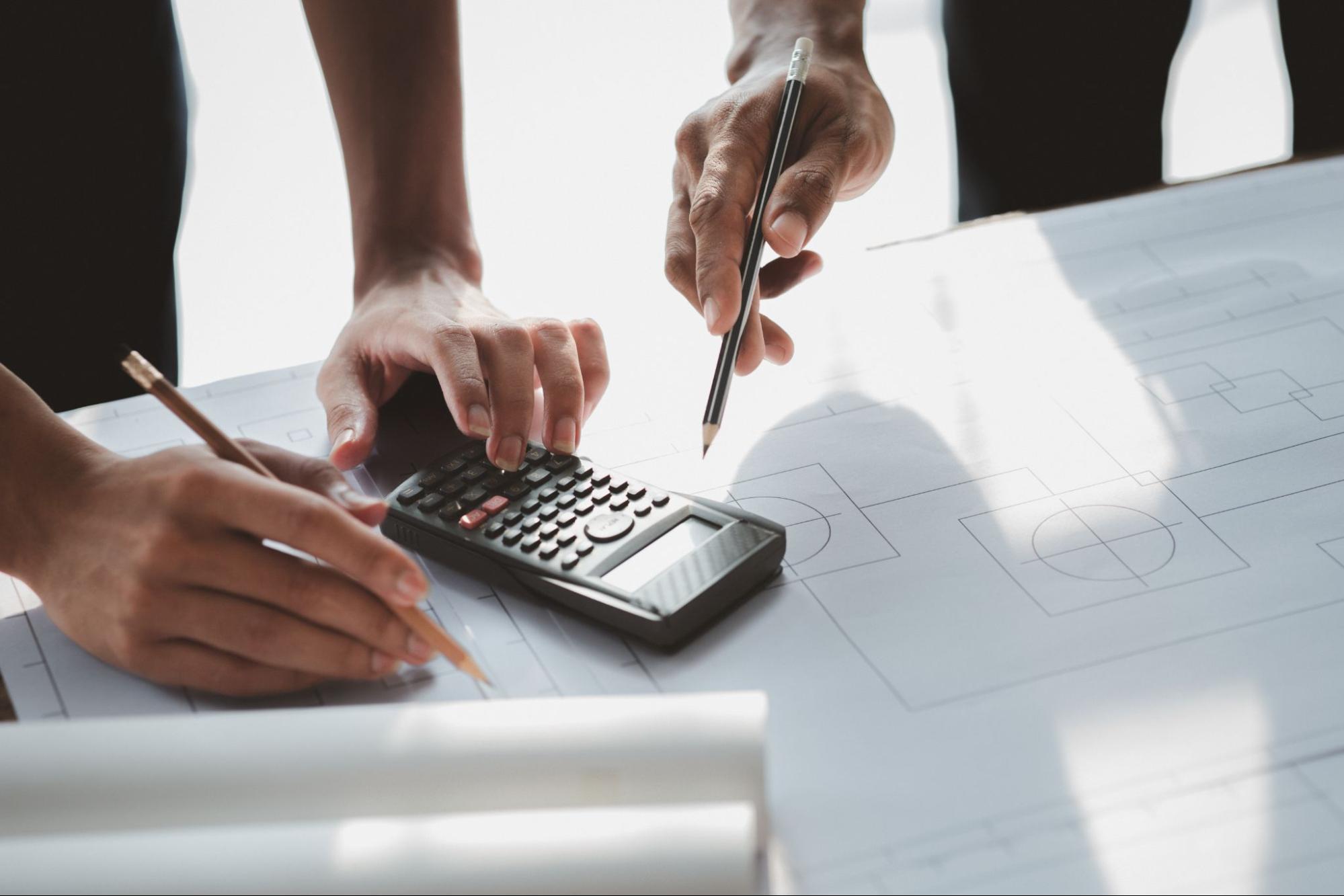 Architects’ hands holding pencils collaborate to adjust a house plan draft on paper, aligning design changes with the project budget requirements.