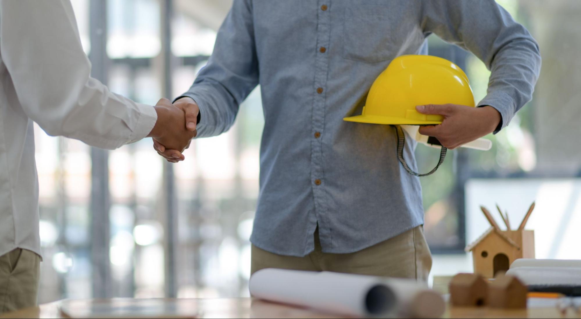 A close-up of a handshake between a business owner and consultant, with one holding a yellow hard hat, symbolizing a partnership in a construction project.