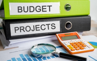 Green binder labeled 'Budget' and black binder labeled 'Project' on a table with financial reports, data charts, a calculator, and a magnifying lens for project cost analysis.