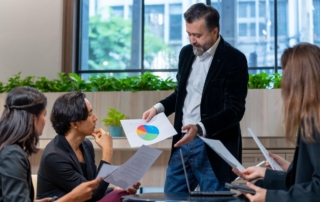 A Caucasian male consultant, standing and holding a paper with a graph, explains data insights to clients in a business meeting.