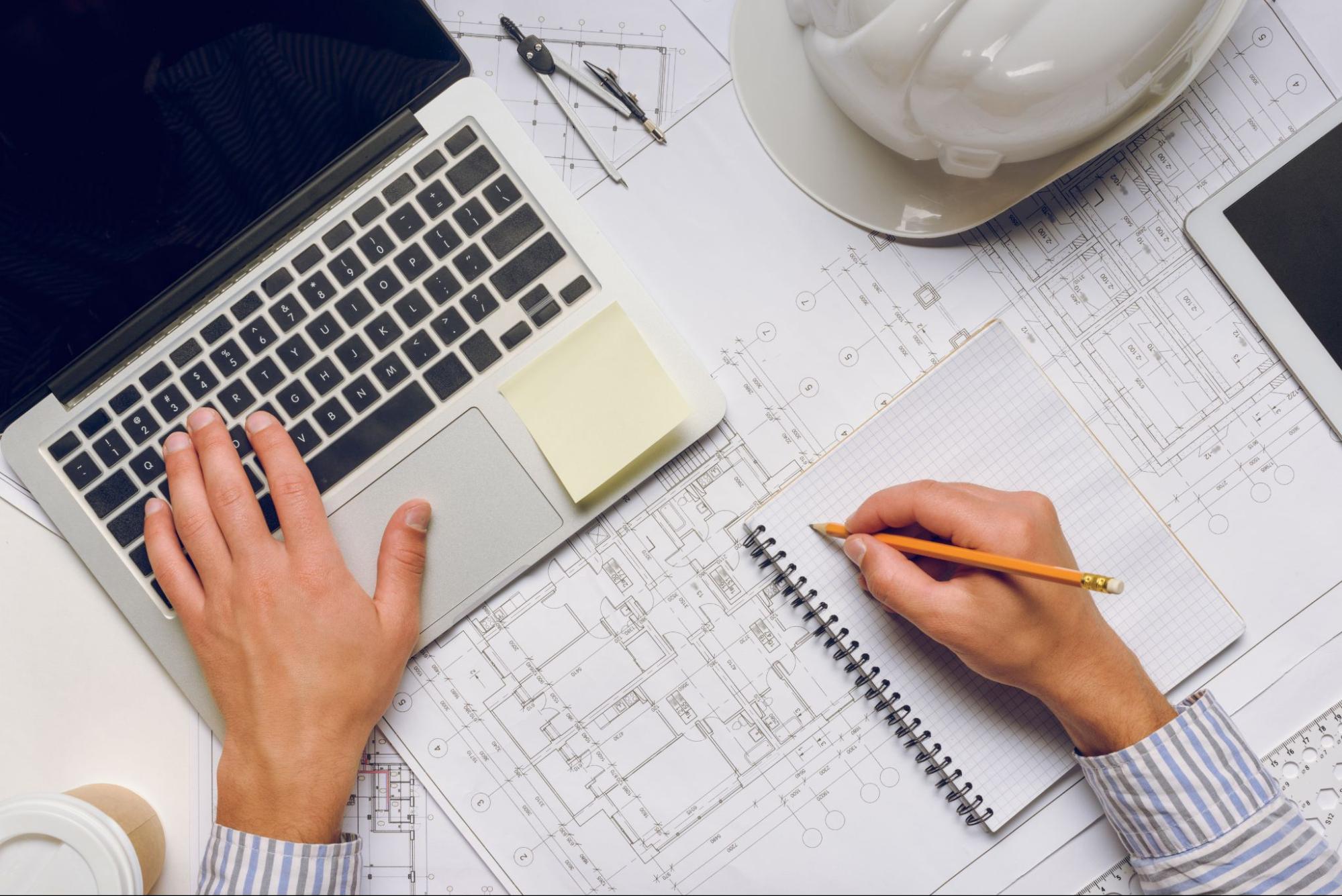 A cropped shot of an architect working on a laptop and writing notes in a notebook, with a blueprint underneath, a compass, a tablet, and a white hard hat on the side.