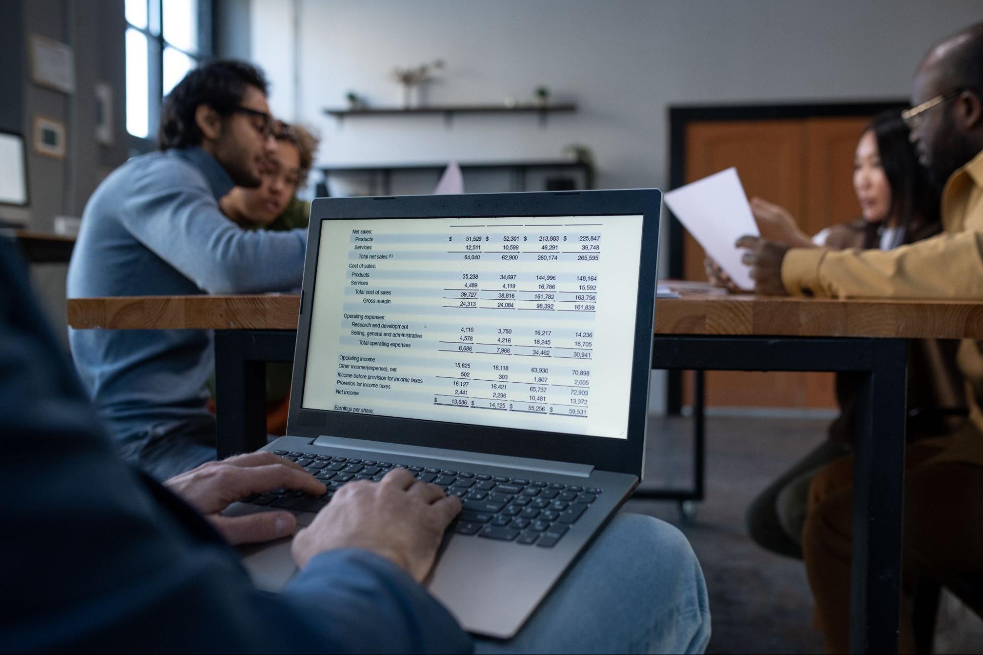 Hands of a businessman typing on a laptop keyboard to prepare a report, with colleagues collaborating in the background.